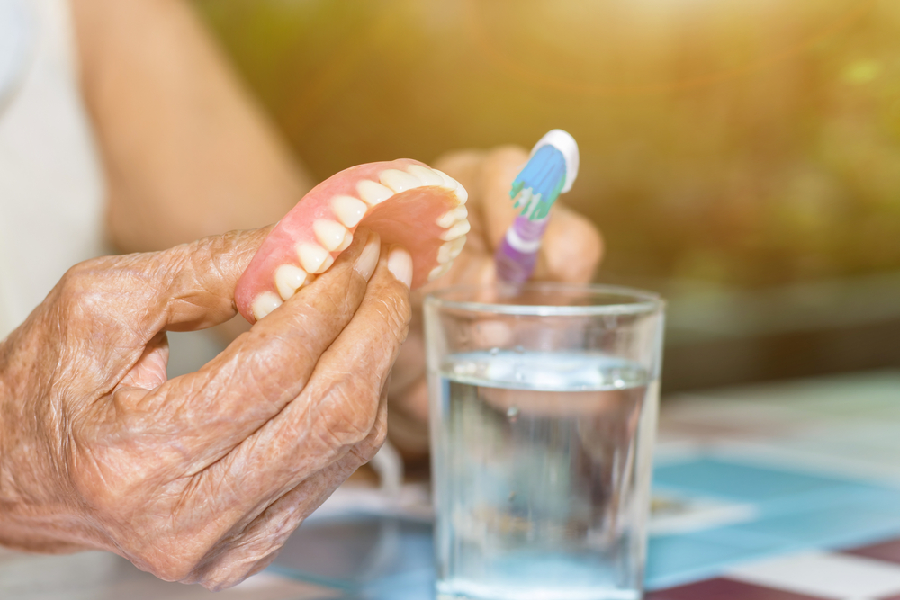 Elderly person holding dentures and toothbrush near a glass of water, emphasizing denture care and maintenance for oral health.