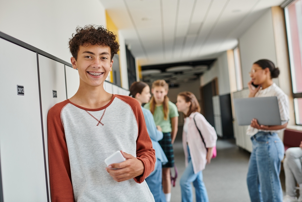 Happy,Boy,With,Braces,Holding,Smartphone,And,Looking,At,Camera Teen boy with braces smiling in school hallway, surrounded by peers discussing and using technology, representing youthful orthodontic care and social interactions.